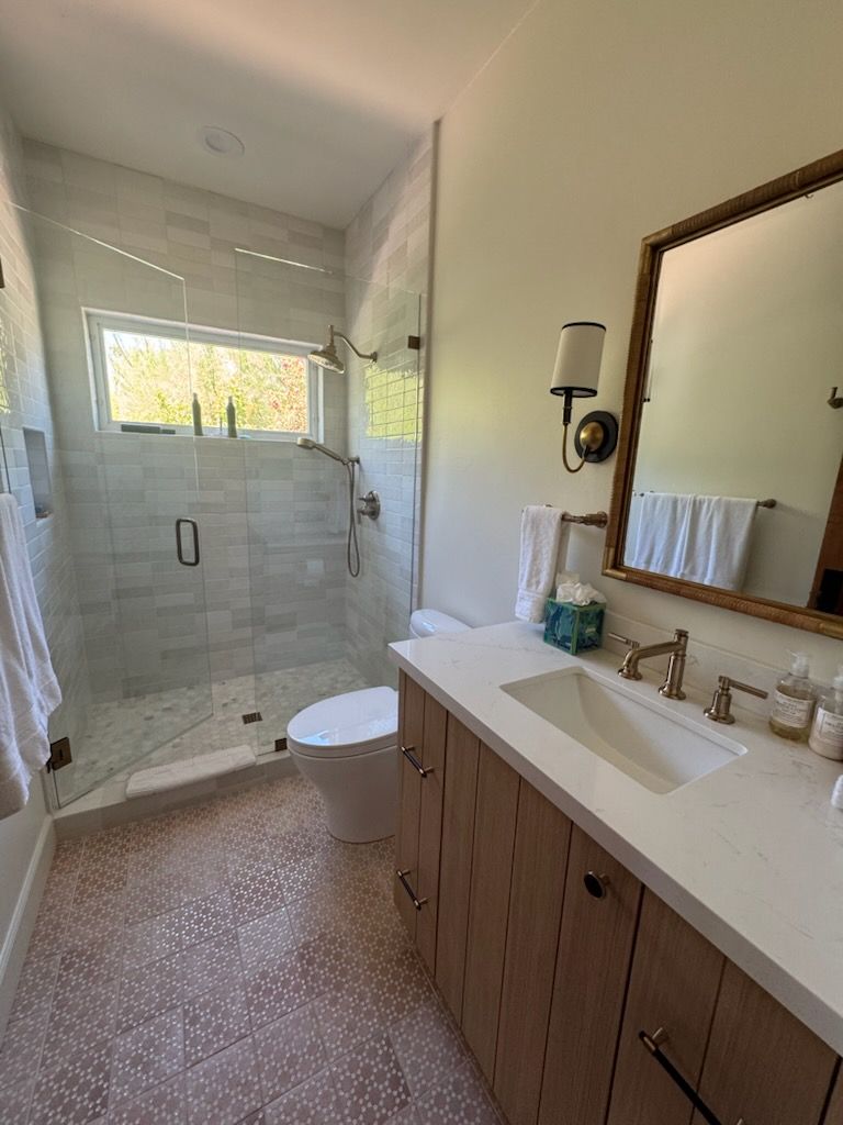 Bathroom with wood vanity, patterned pink tile floor, glass shower, gold fixtures, and window.