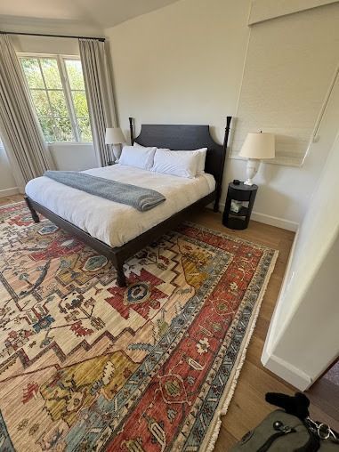 Bedroom with bed, nightstand, lamps, patterned rug, and window with curtains.