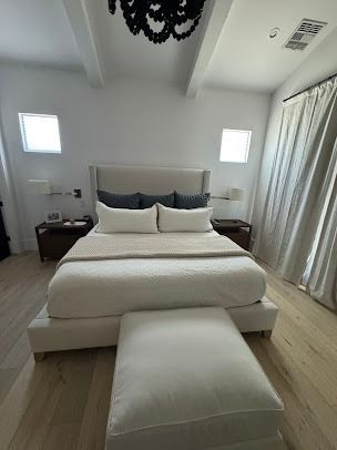 A bright bedroom with a large bed, nightstands, and an ottoman. Light-colored walls, floors, and curtains.