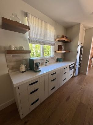 Kitchen with white cabinets, black handles, white countertops, open shelves, and a window with a shade.