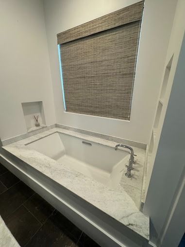 Modern bathroom with white tub set in marble, window with brown shade, built-in shelves, and dark wood floor.