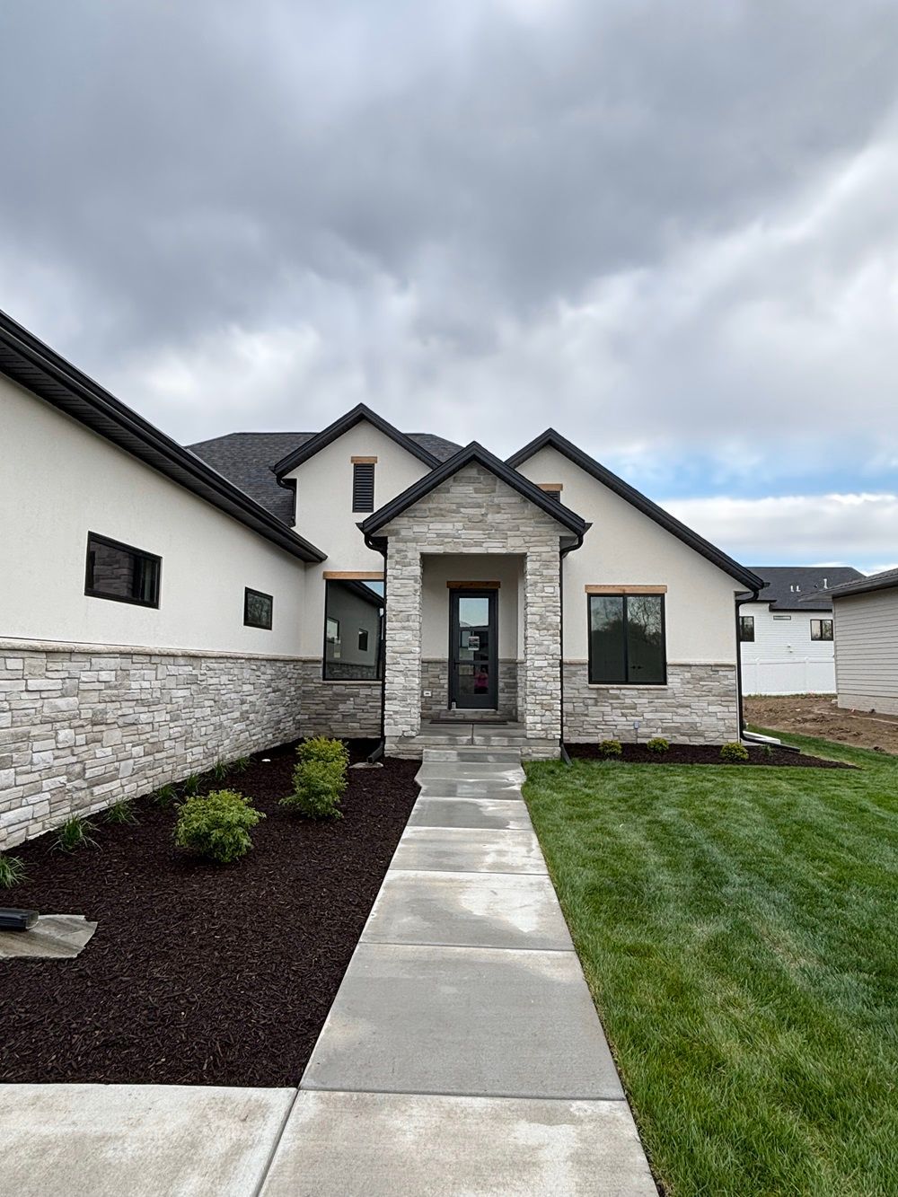 Modern home with gray stone and white stucco exterior, concrete walkway, and manicured lawn under overcast sky.