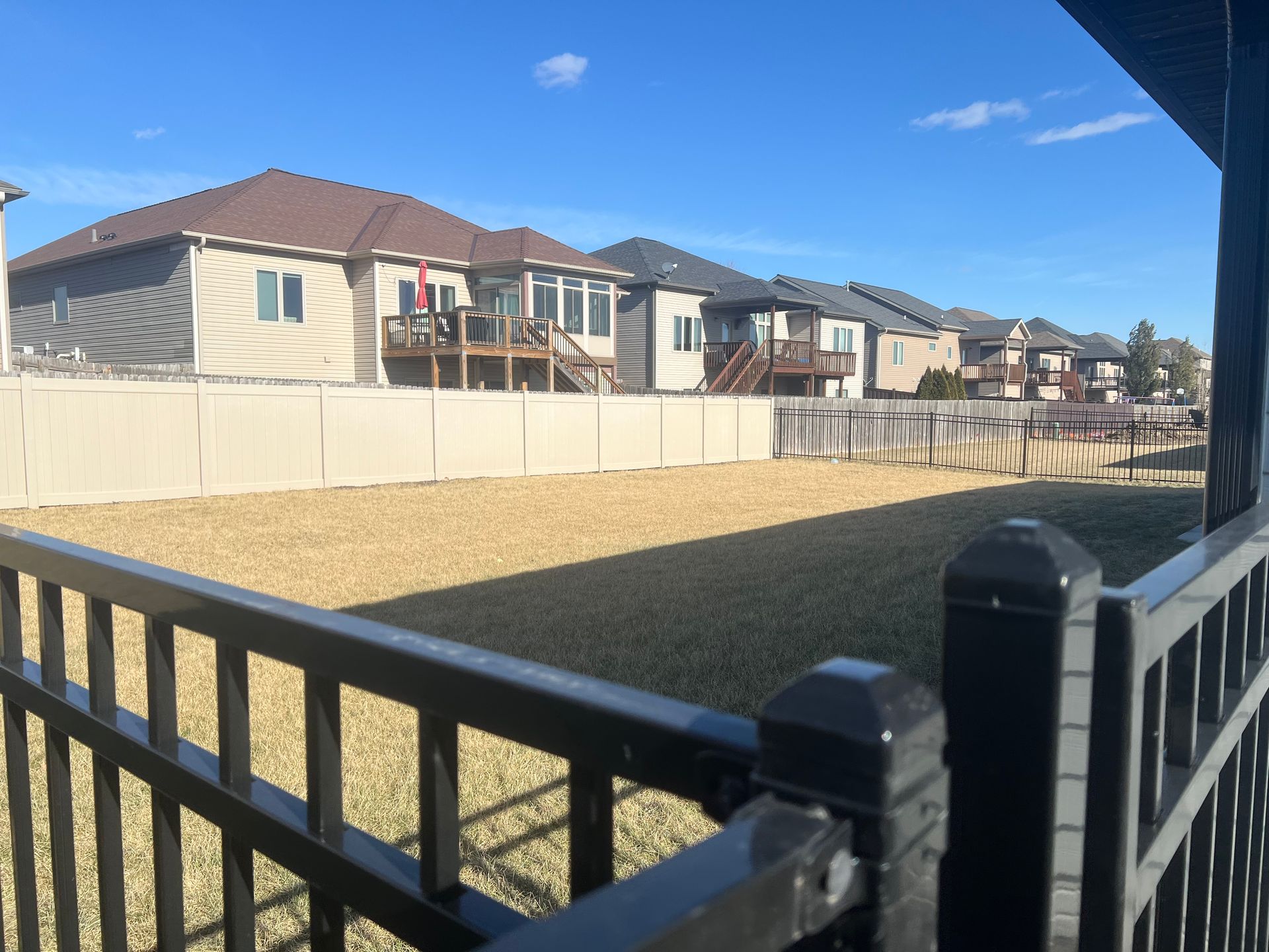 View of a backyard with a fence, and homes under a bright blue sky.