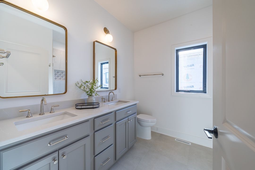 Bathroom with gray vanity, two gold-framed mirrors, white walls, and a window.