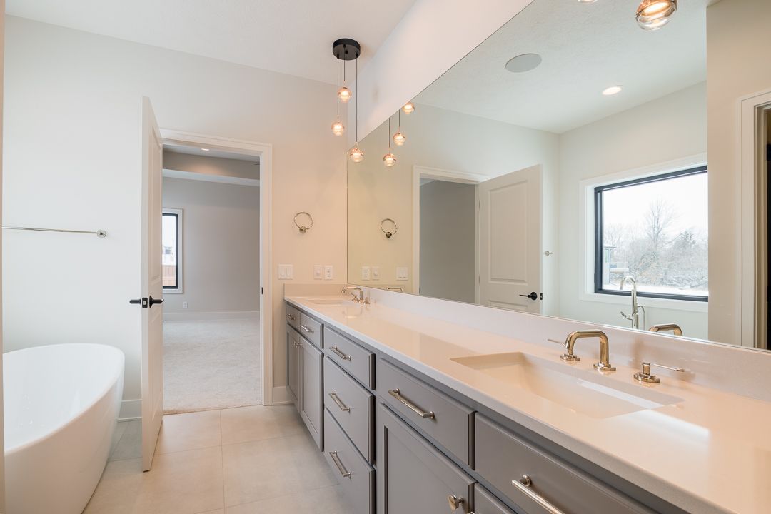 Spacious bathroom with gray cabinets, large mirror, and soaking tub. Light fixture and window visible.