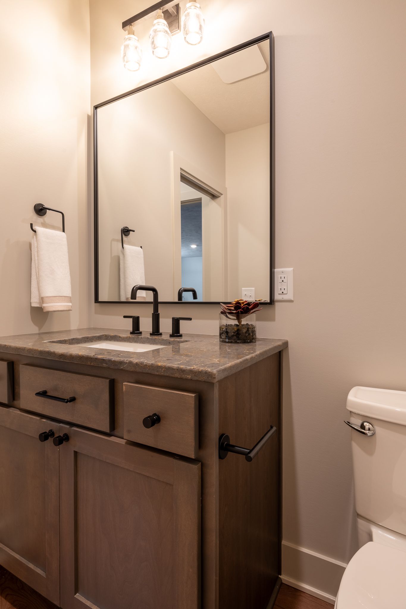 Powder room with a vanity, mirror, and toilet. Features dark fixtures and neutral walls.