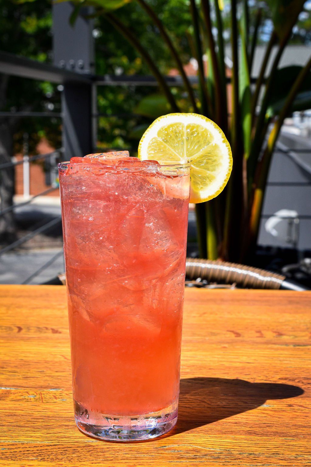 a tall glass of pink liquid with ice and a lemon slice on a wooden table .