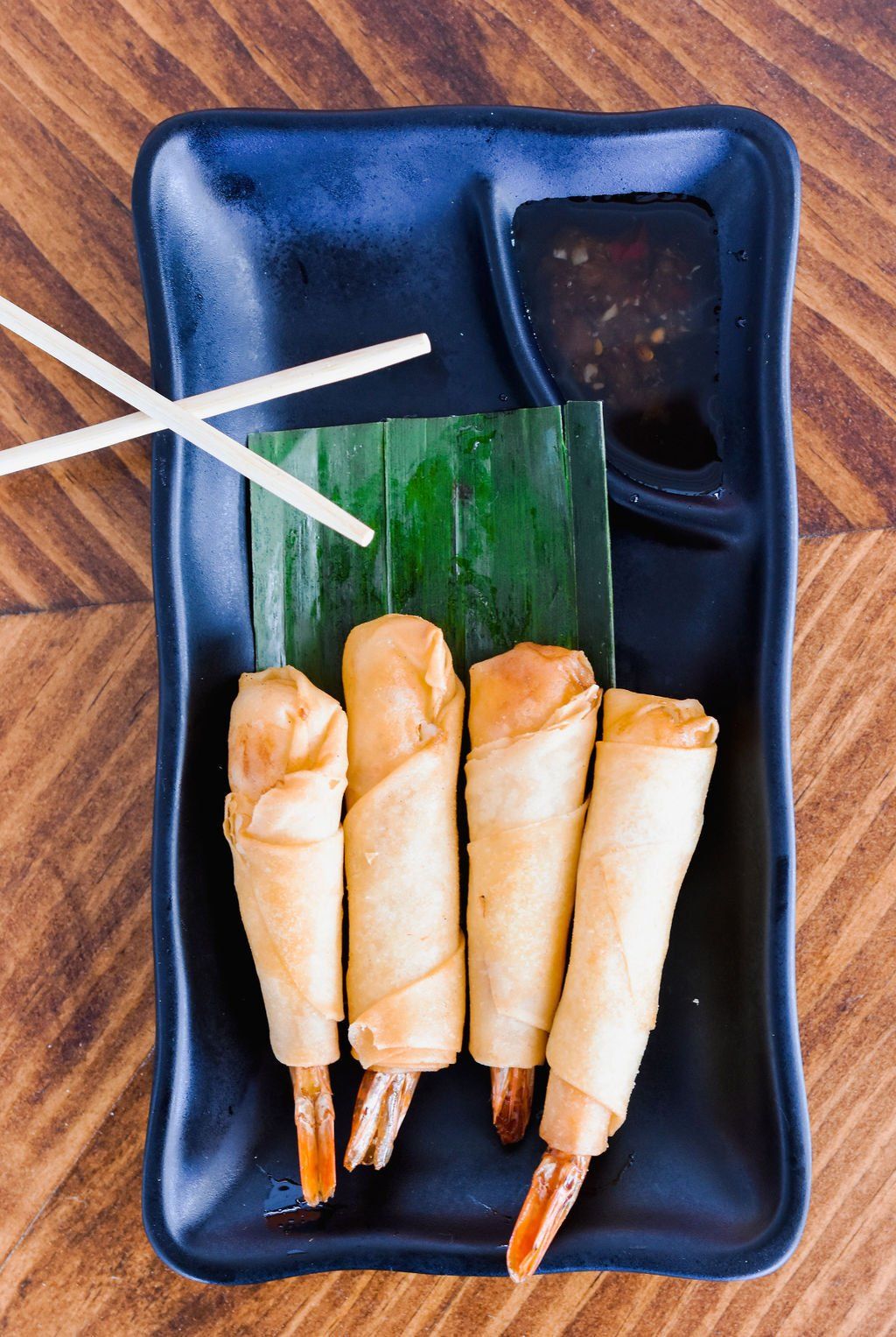 a black plate topped with shrimp rolls and chopsticks on a wooden table .