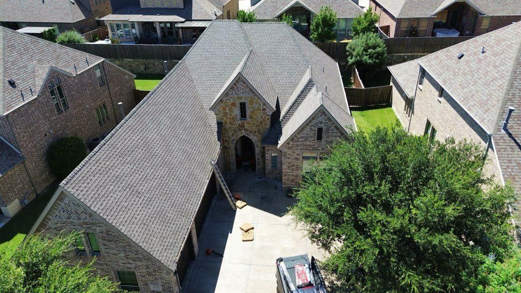 Aerial view of a brown stone house with a complex roof structure and a driveway; surrounded by trees and other houses.
