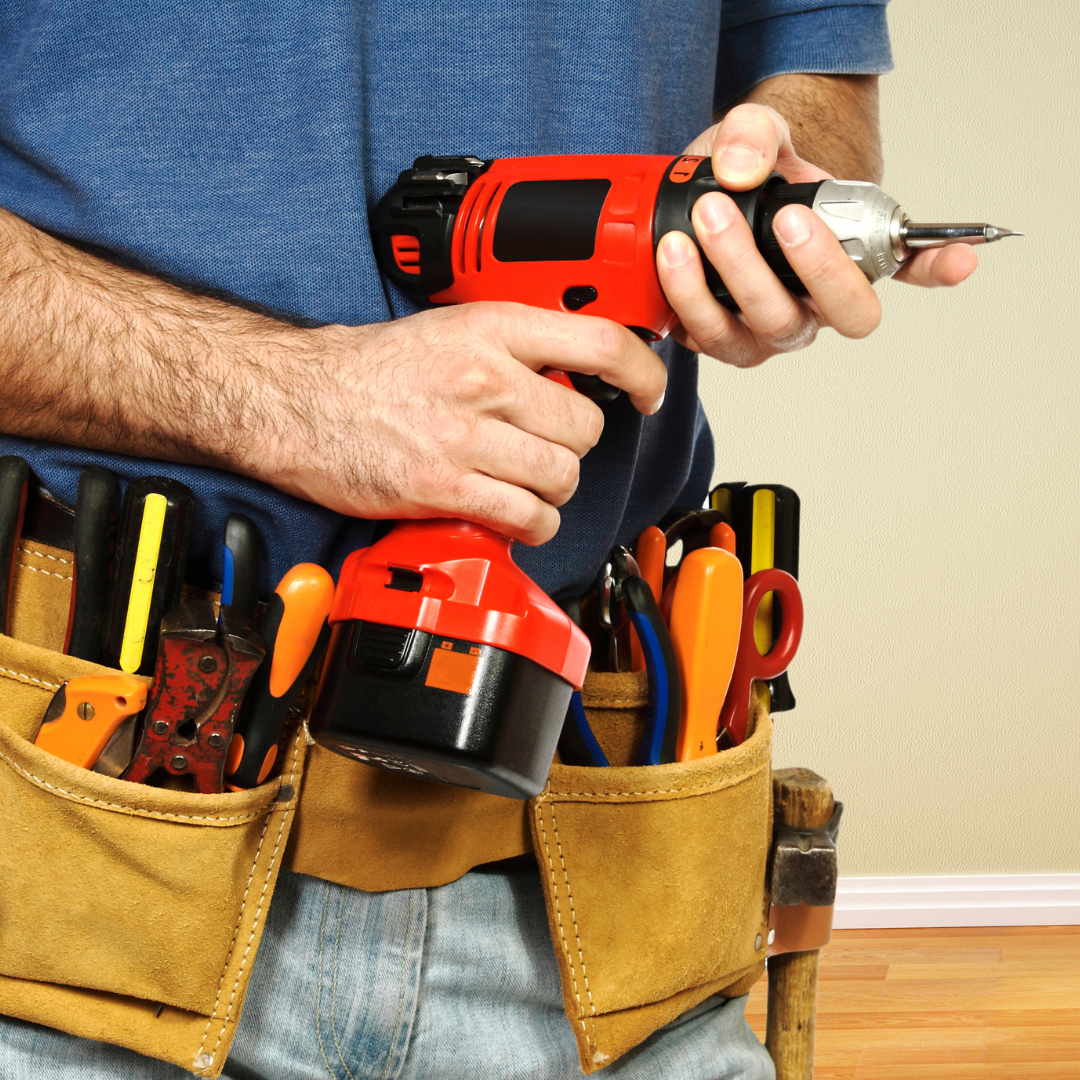 Man holding a red drill, wearing a tool belt with various tools.