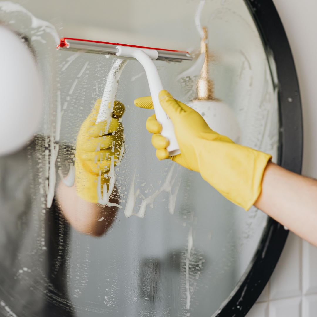 Person in yellow gloves uses a squeegee to clean a round bathroom mirror.