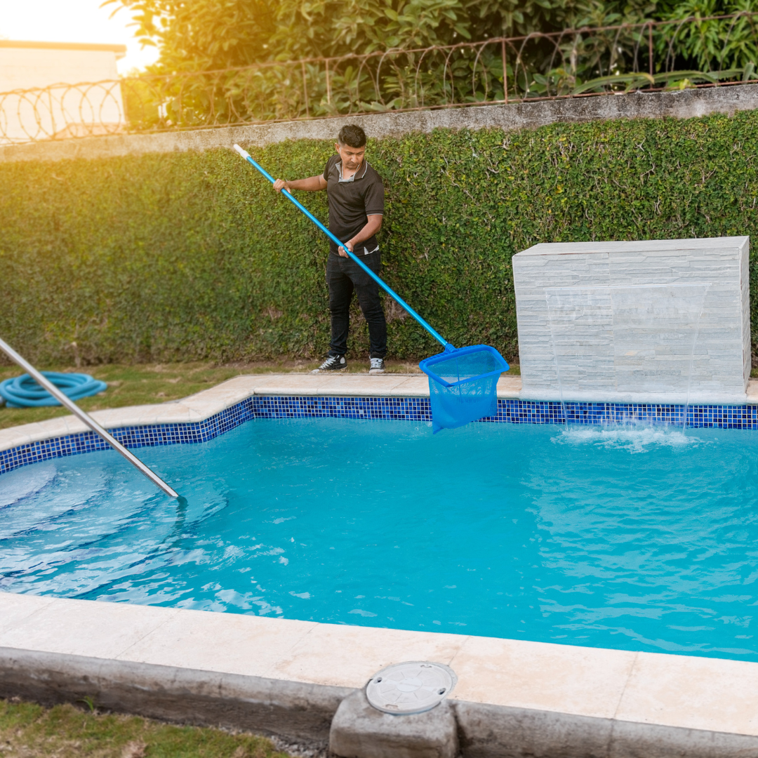 Person cleaning a blue pool with a net. Lush green yard in the background.