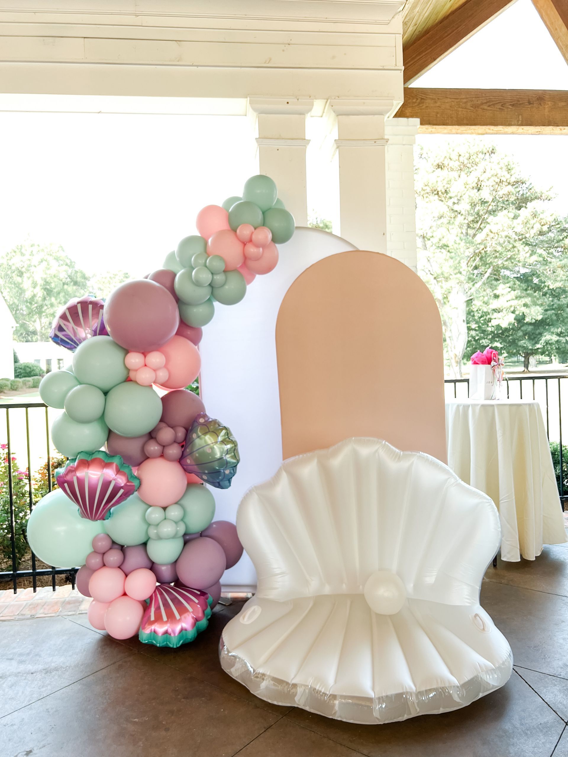 A mermaid-themed event display featuring a large white shell, a peach-colored arched panel, and a balloon garland.