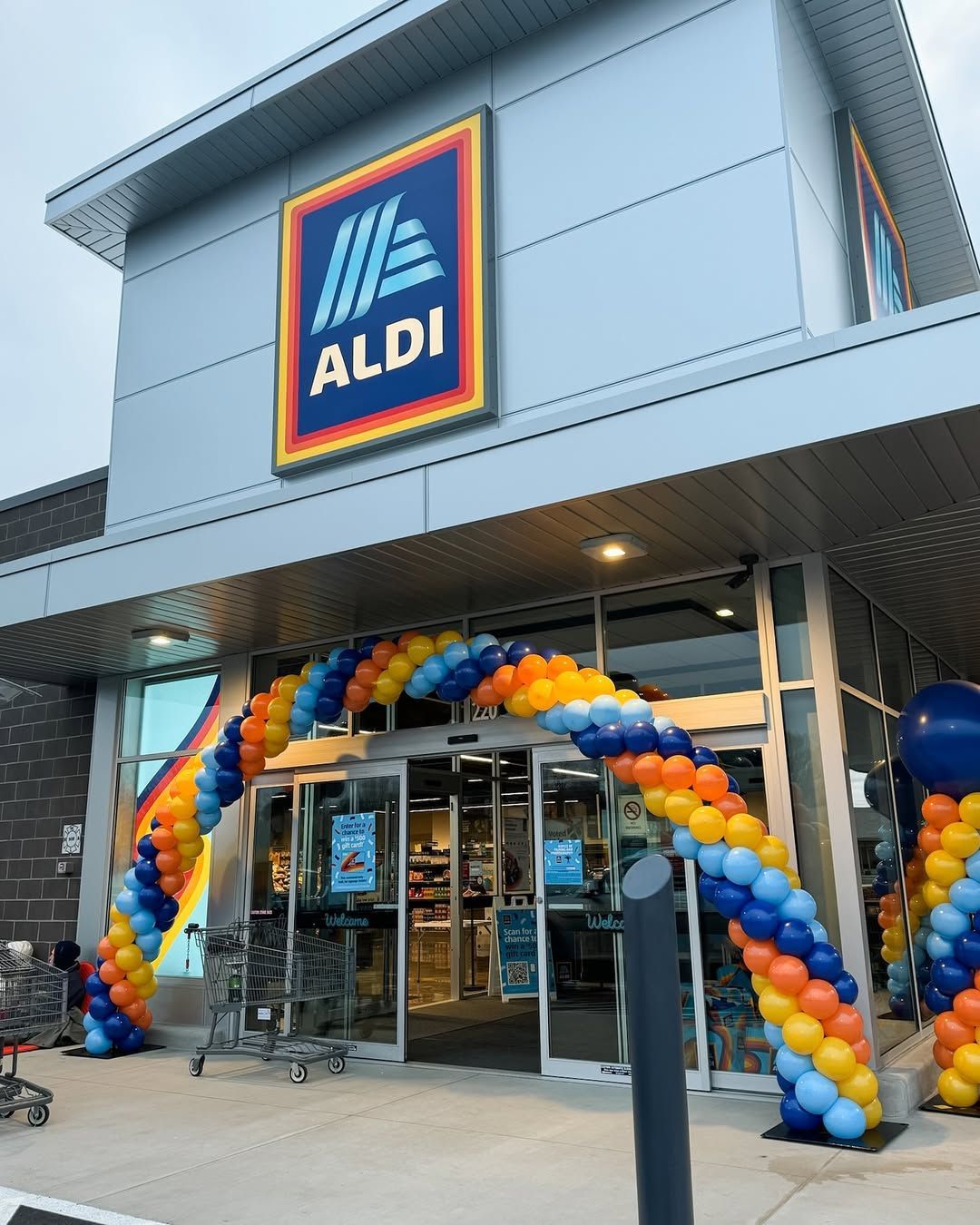 The exterior of an Aldi grocery store with a celebratory balloon arch in blue, orange, and yellow over the entrance.