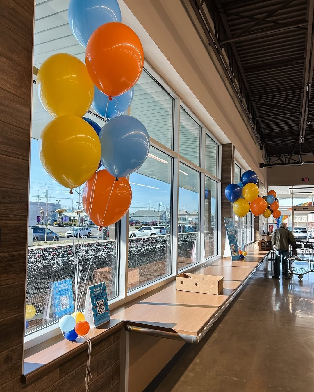 Clusters of blue, yellow, and orange balloons decorate windows inside a store with an open counter and view of a parking lot.