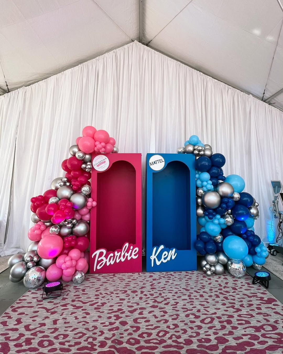 A party backdrop with two life-sized doll boxes in pink and blue, decorated with matching balloon arches on a rug.