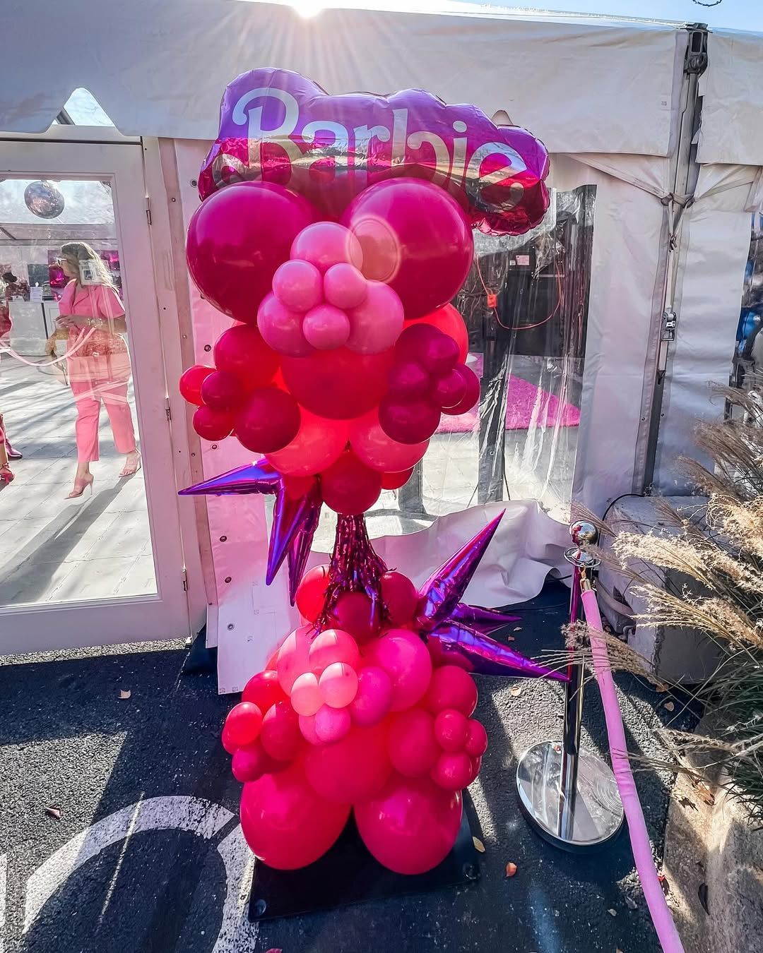 A Barbie-themed balloon arch stands on a patio, featuring shades of pink balloons and a Barbie logo foil balloon topper.