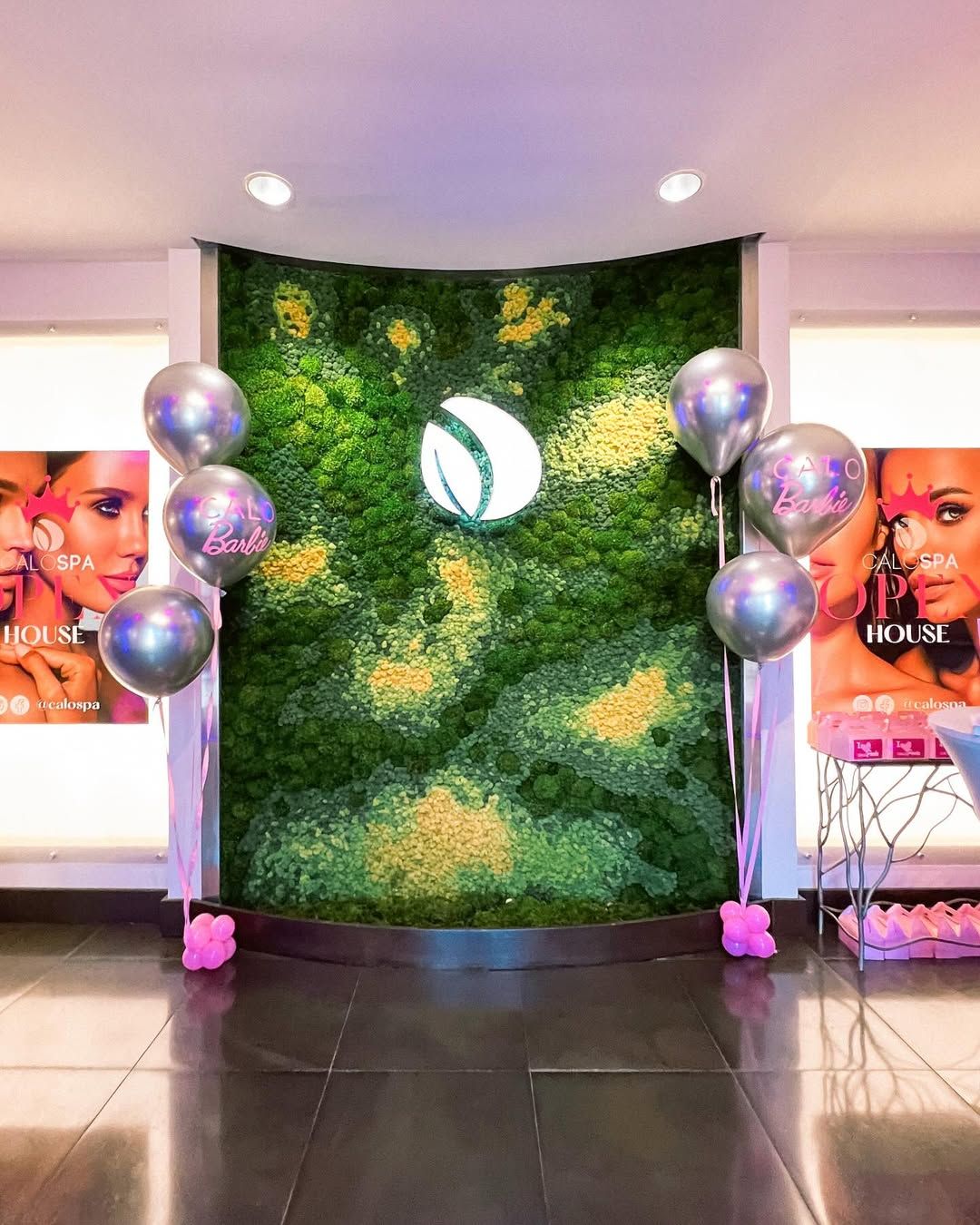 A moss wall with a backlit circular logo, flanked by two balloon clusters, set between beauty spa advertising posters.