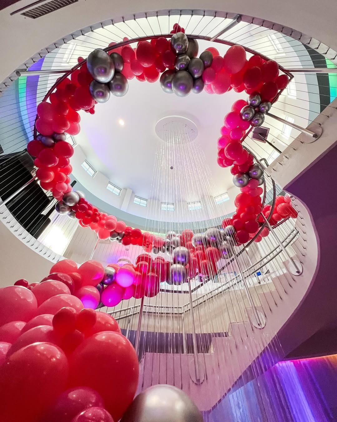 A winding staircase is decorated with a vibrant arch of red and silver balloons, with cascading light fixtures above.