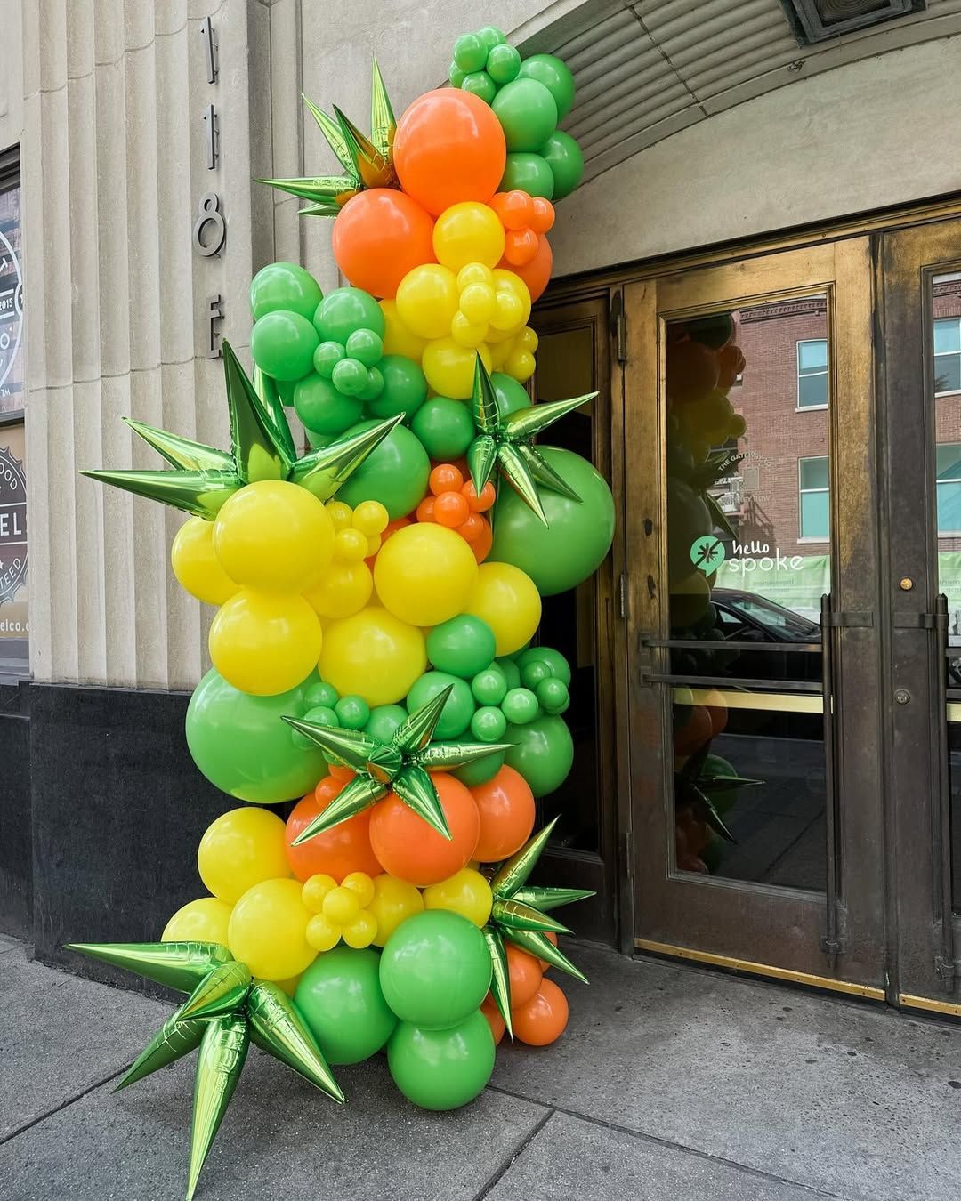 A vertical balloon arch standing by an entrance, featuring clusters of green, yellow, and orange balloons with star accents.