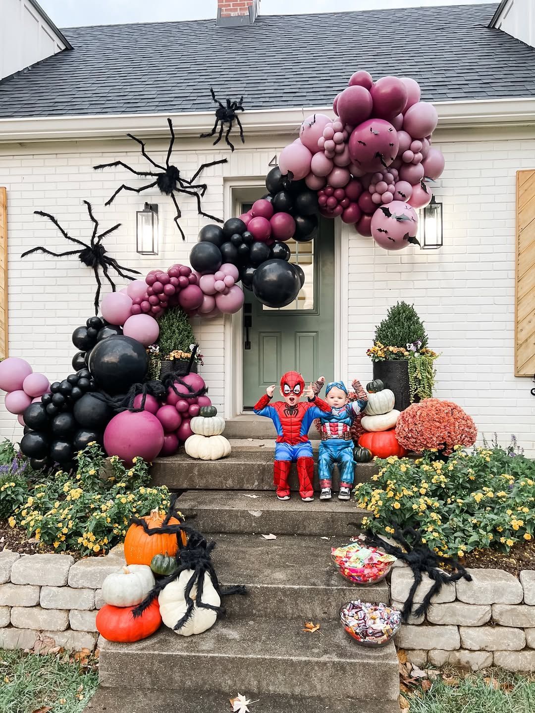 Two children in costumes sit on steps in front of a white house decorated with a purple and black Halloween balloon arch.