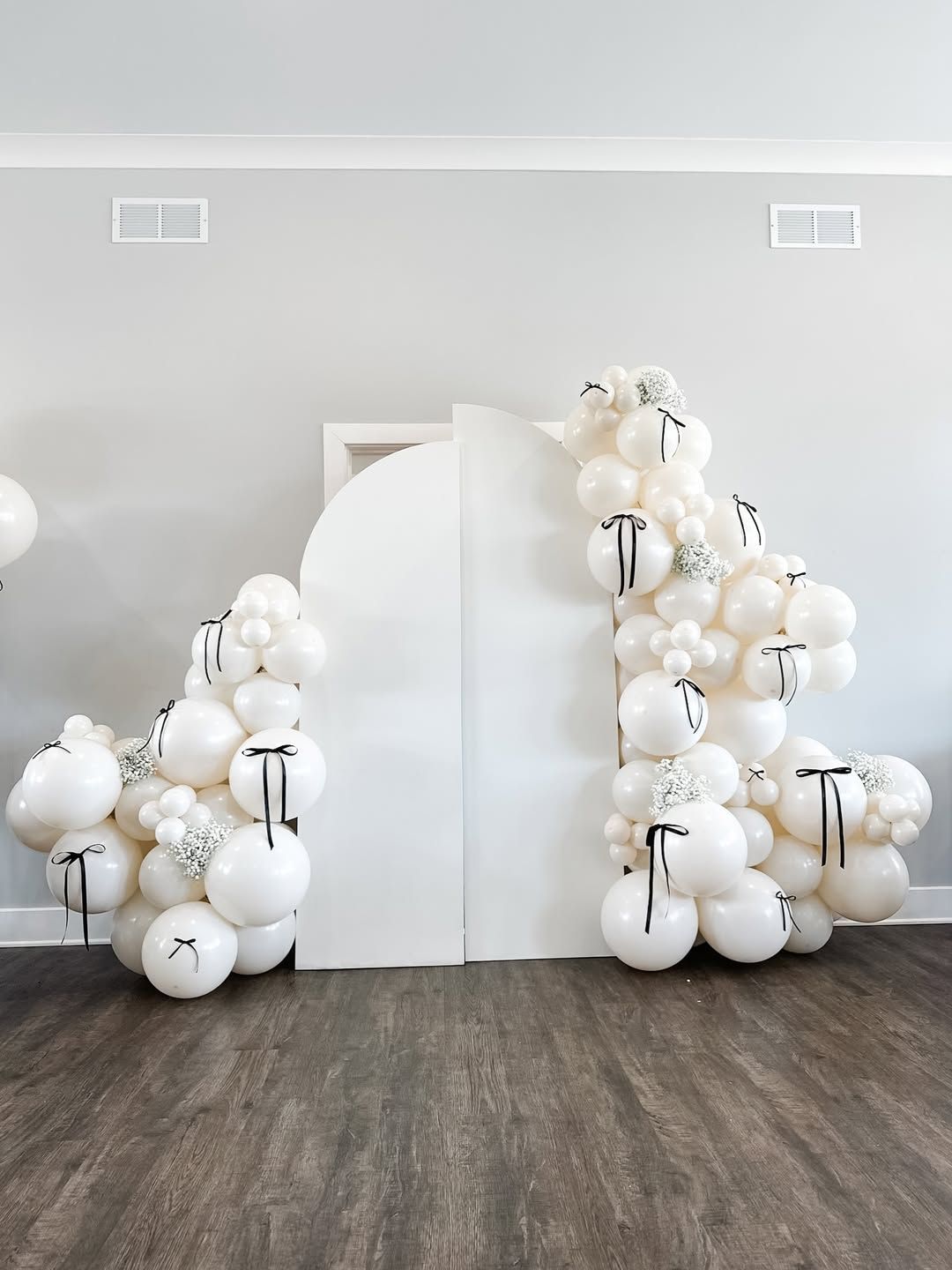 A bridal shower sign with a white bow stands next to white balloon bunches and a blue armchair in a brightly lit room.