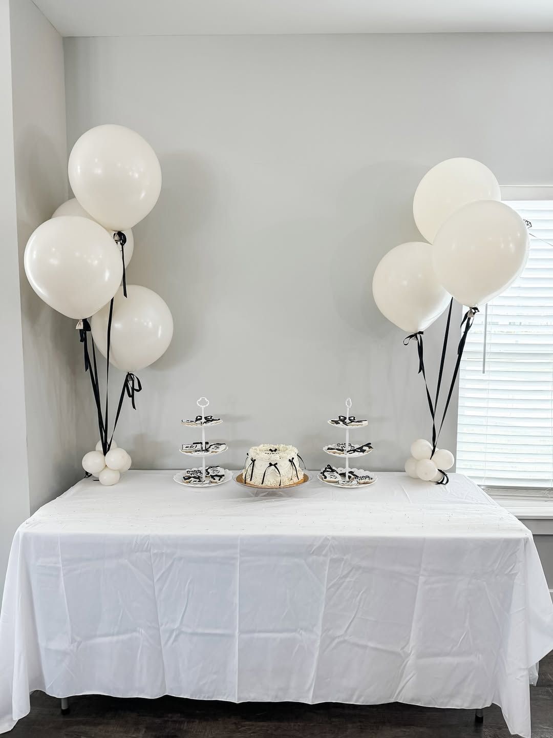 A white-clothed dessert table features two balloon clusters, two small three-tier stands, and a centered bundt cake.