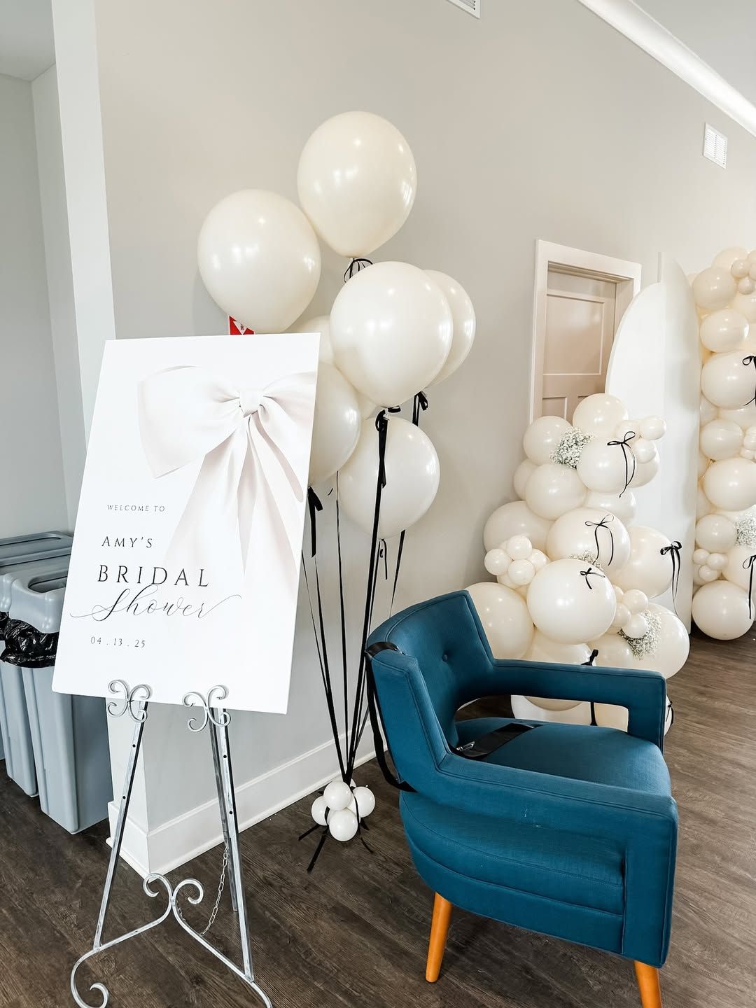 A white arched backdrop decorated with white balloons and small black ribbons, set against a wall on a wooden floor.