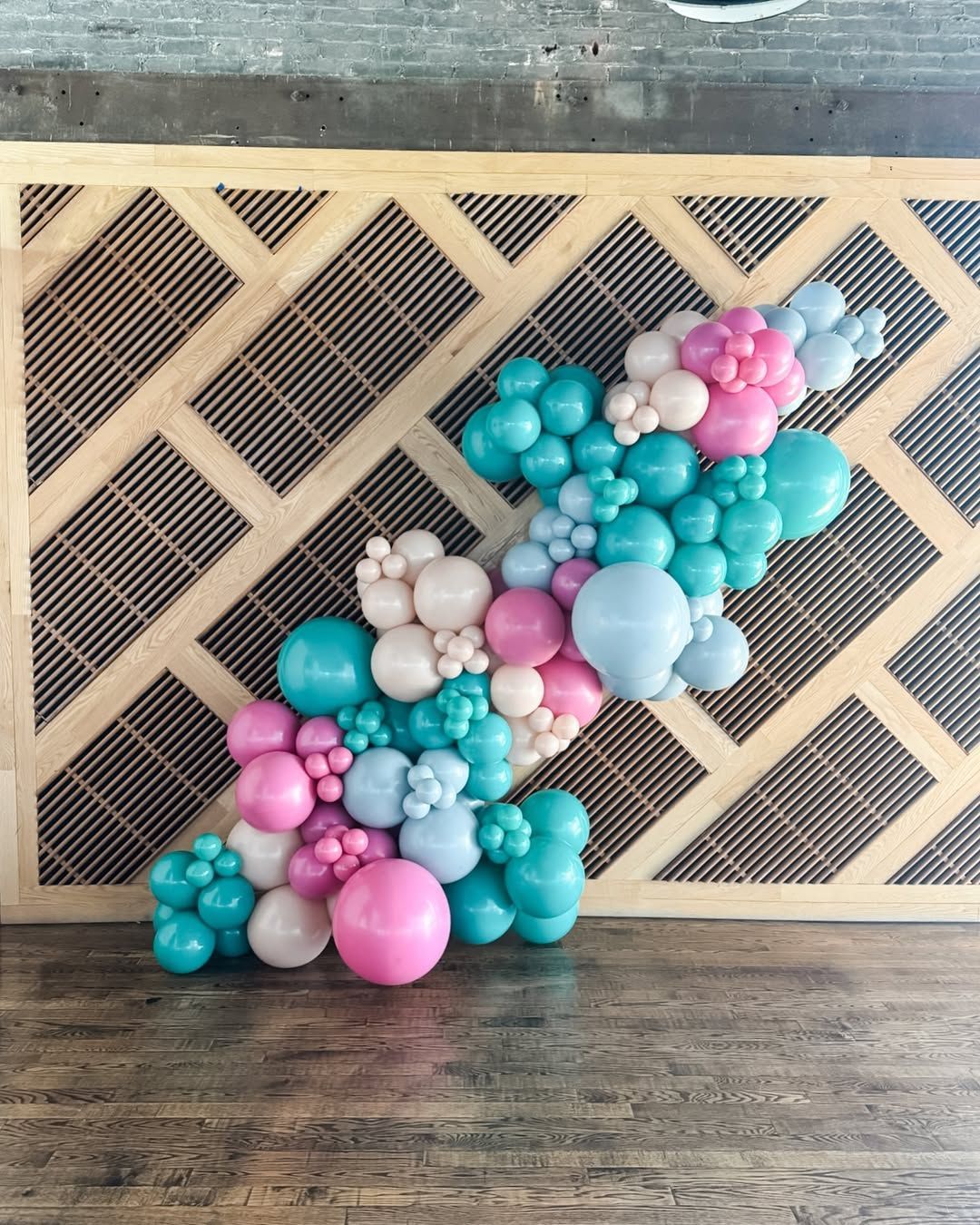 A balloon garland featuring varying sizes of teal, light blue, and pink balloons mounted on a tan lattice-style wall.