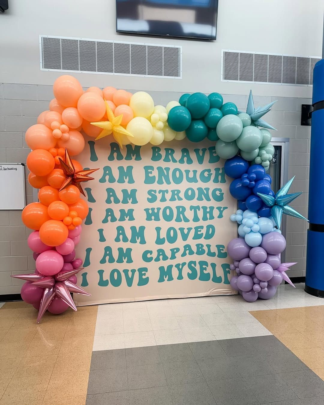 A backdrop with encouraging affirmations in teal text, surrounded by a colorful balloon arch, in a building interior.