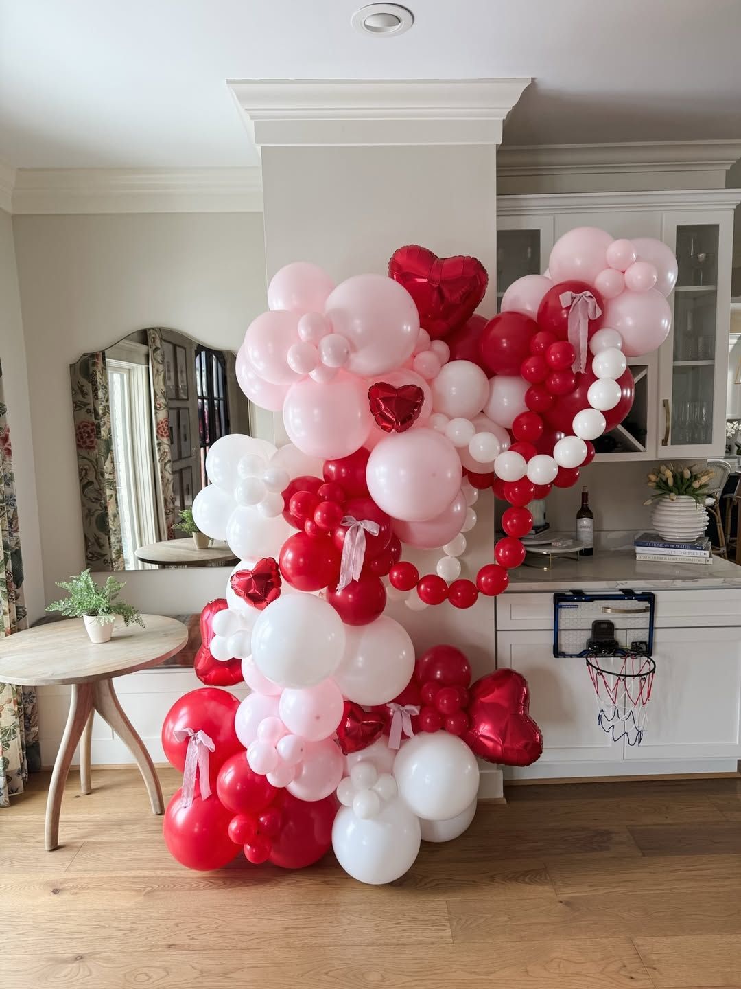 A large, vibrant balloon garland in red, white, and pink shades, featuring heart-shaped accents in an indoor home setting.