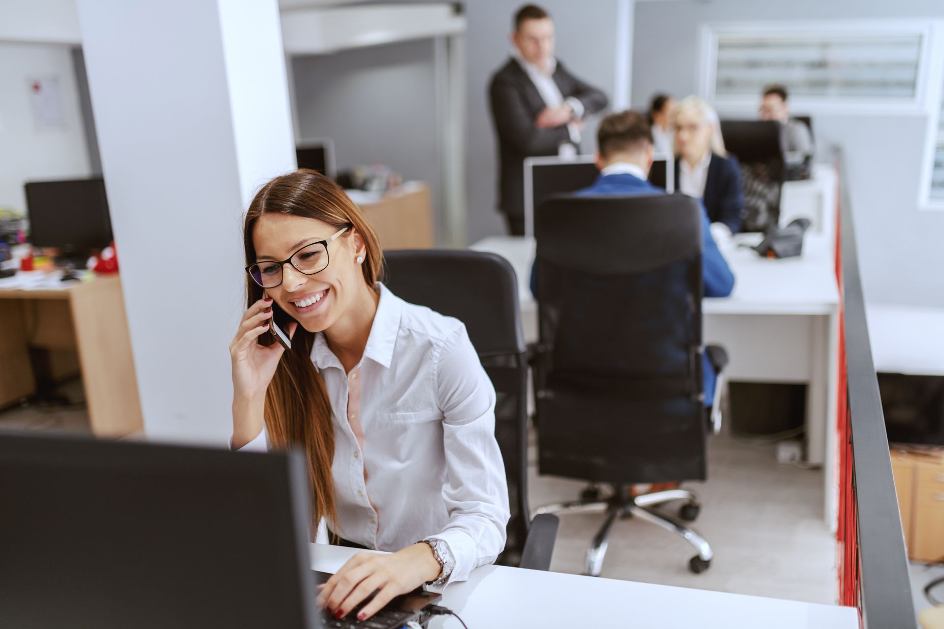 A woman is using an office computer and talking on the phone. Her coworkers are in the background.