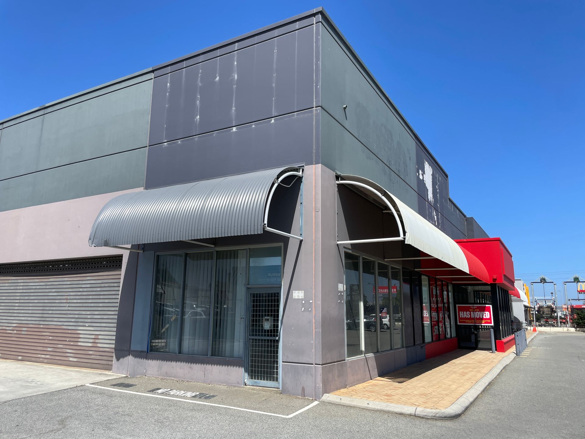 Exterior of a commercial building with dark gray and red accents, awnings, and storefront windows under a clear, blue sky.