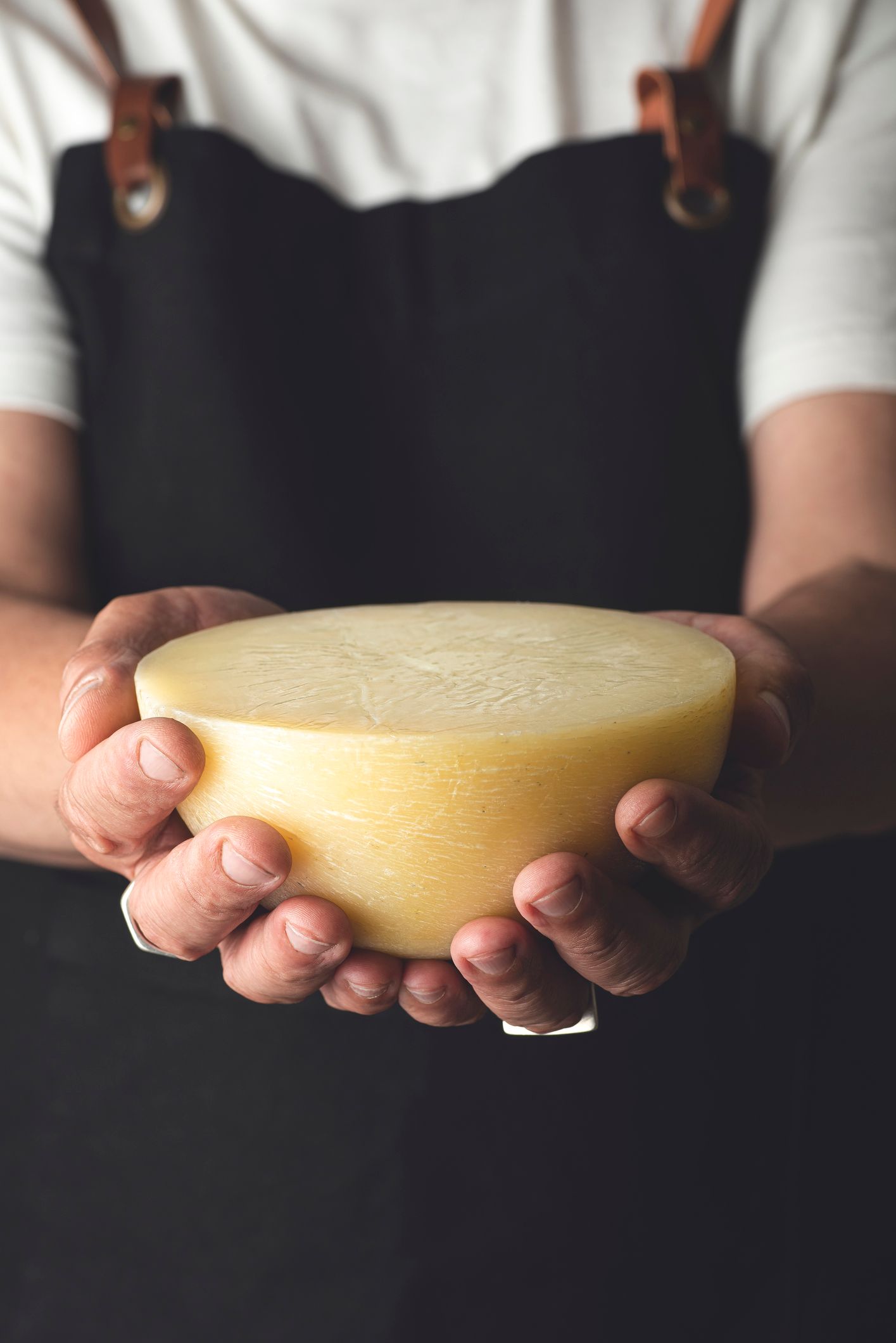 Persona che tiene in entrambe le mani un formaggio rotondo e giallo e indossa un grembiule nero.