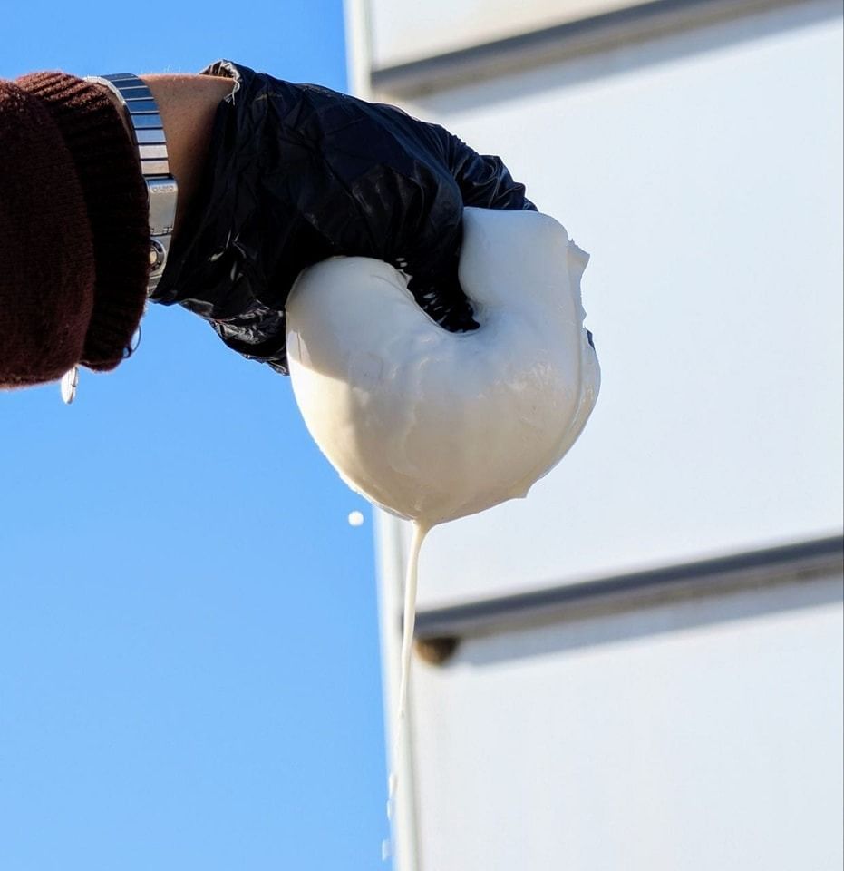 Una persona con un guanto nero spreme una sostanza bianca, simile a un impasto; il liquido gocciola. Sullo sfondo, cielo e superficie bianca.