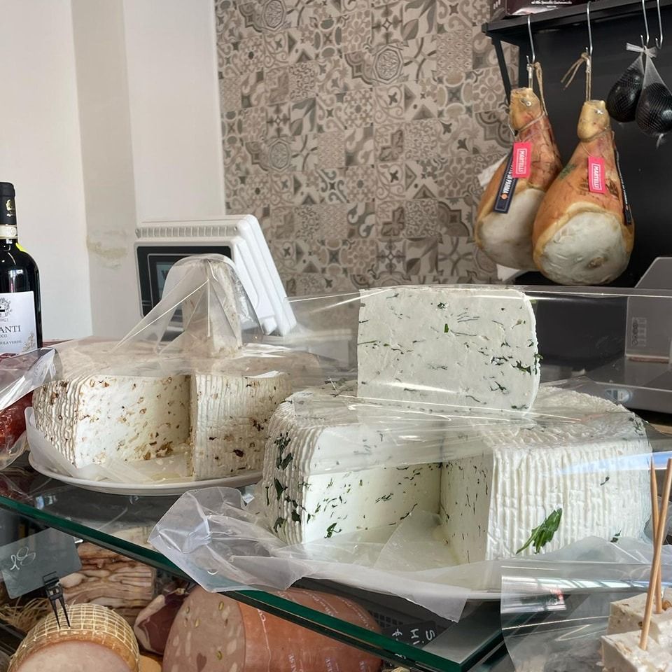 Esposizione di formaggi in una gastronomia, con forme incartate e salumi appesi sullo sfondo.