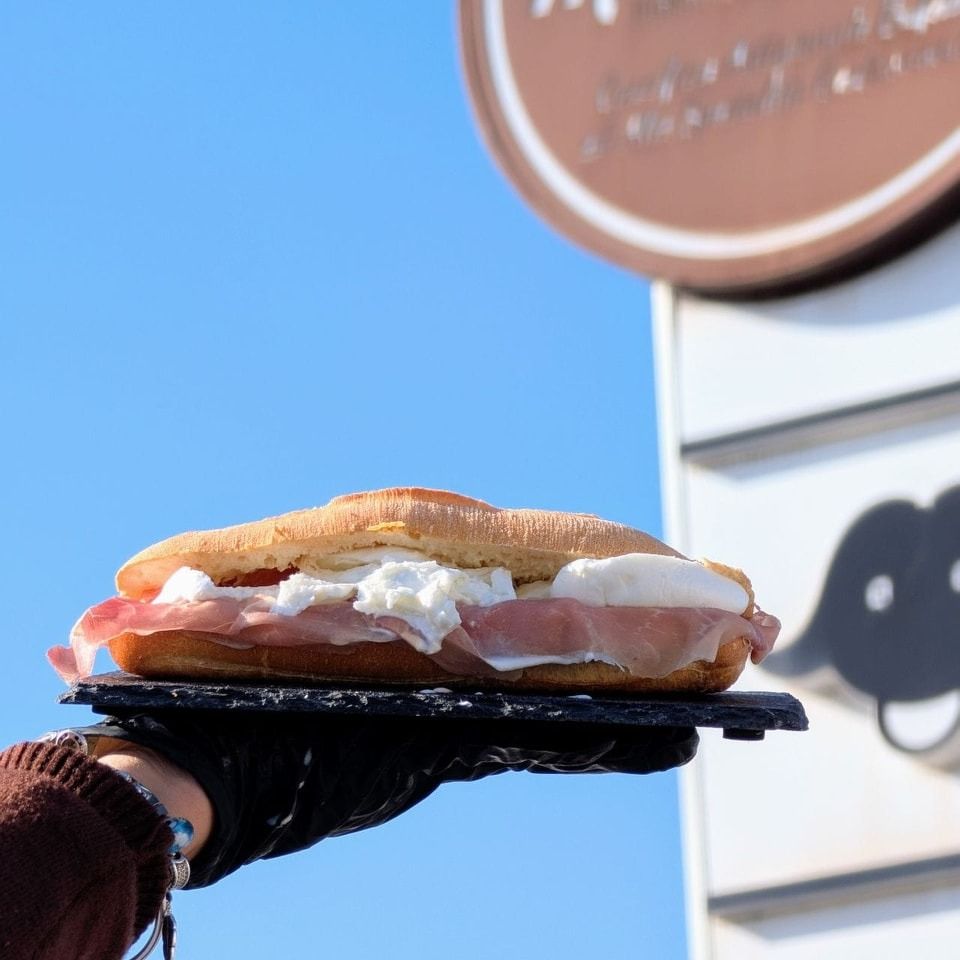 Mano che tiene un panino con prosciutto e formaggio cremoso su uno sfondo di ardesia nera e cielo azzurro.