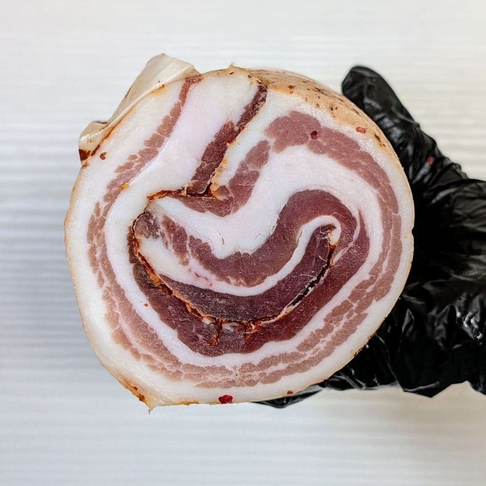 Sezione trasversale di un salume arrotolato, con strati alternati di carne rossa e grasso bianco, tenuto da una mano guantata.