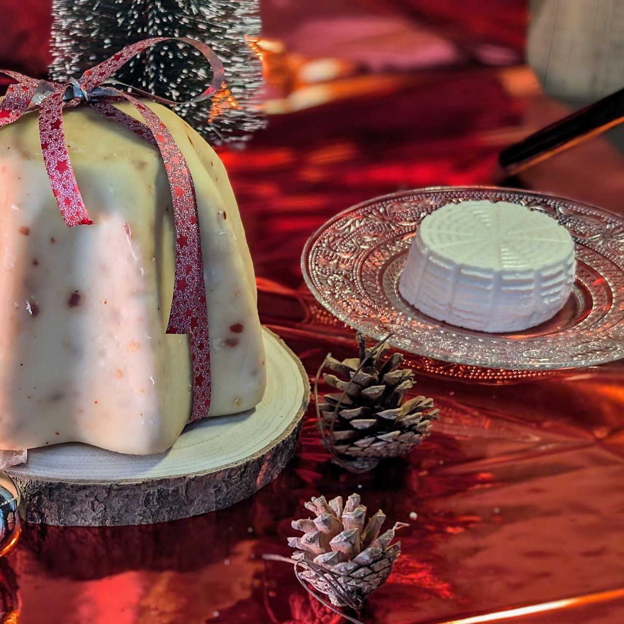 Scena di dolce natalizio: panettone con nastro su tavola di legno, ricotta, pigne su una superficie rossa.