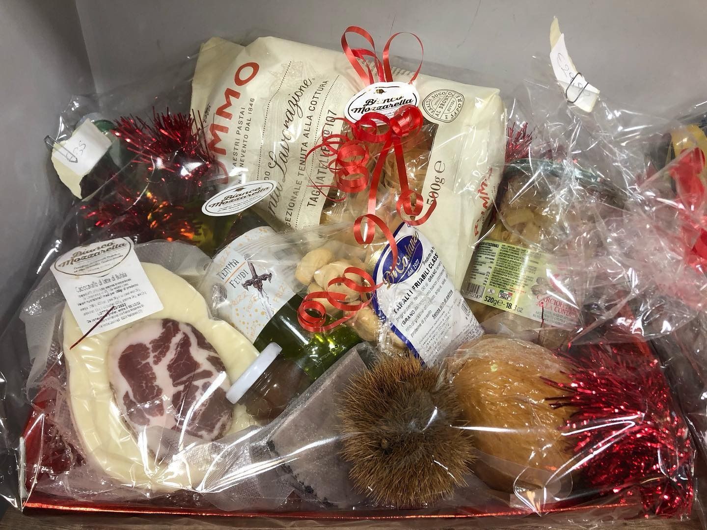 Cesto regalo pieno di prodotti alimentari, avvolto in cellophane e decorato con nastri rossi.