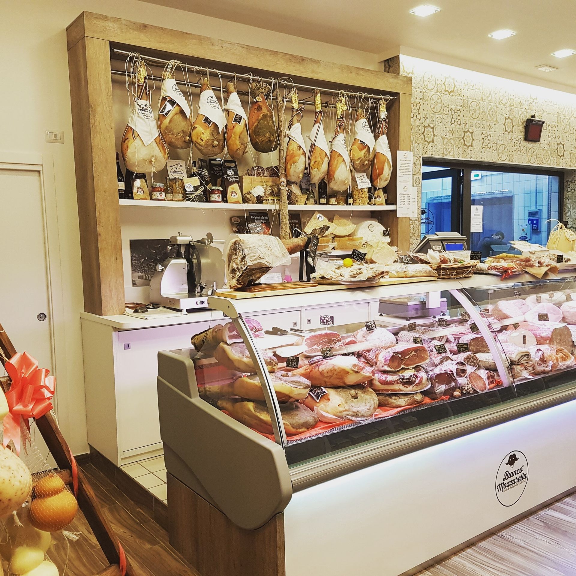 Un banco gastronomia con salumi appesi, un'esposizione refrigerata di carni e scaffali di prodotti alimentari.