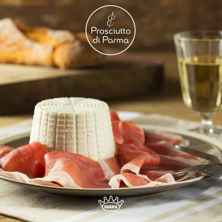 Prosciutto di Parma con ricotta su un piatto, bicchiere di vino e baguette sullo sfondo.