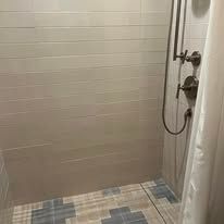 A shower with light gray horizontal subway tiles, a bronze shower head and controls, and a blue-and-white patterned floor.