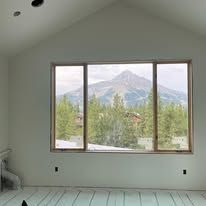 A large window in a room under construction frames a scenic view of a mountain, forest, and sky.