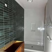 A bathroom with dark green rectangular wall tiles, a wooden vanity, and a glass-enclosed shower with a white tiled interior.