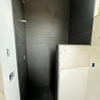 A walk-in shower under construction with dark grey tiled walls, a white half-wall, and exposed wiring on the left.