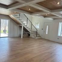 A bright, modern interior room with light wood floors, a coffered wood-paneled ceiling, and a central open staircase.