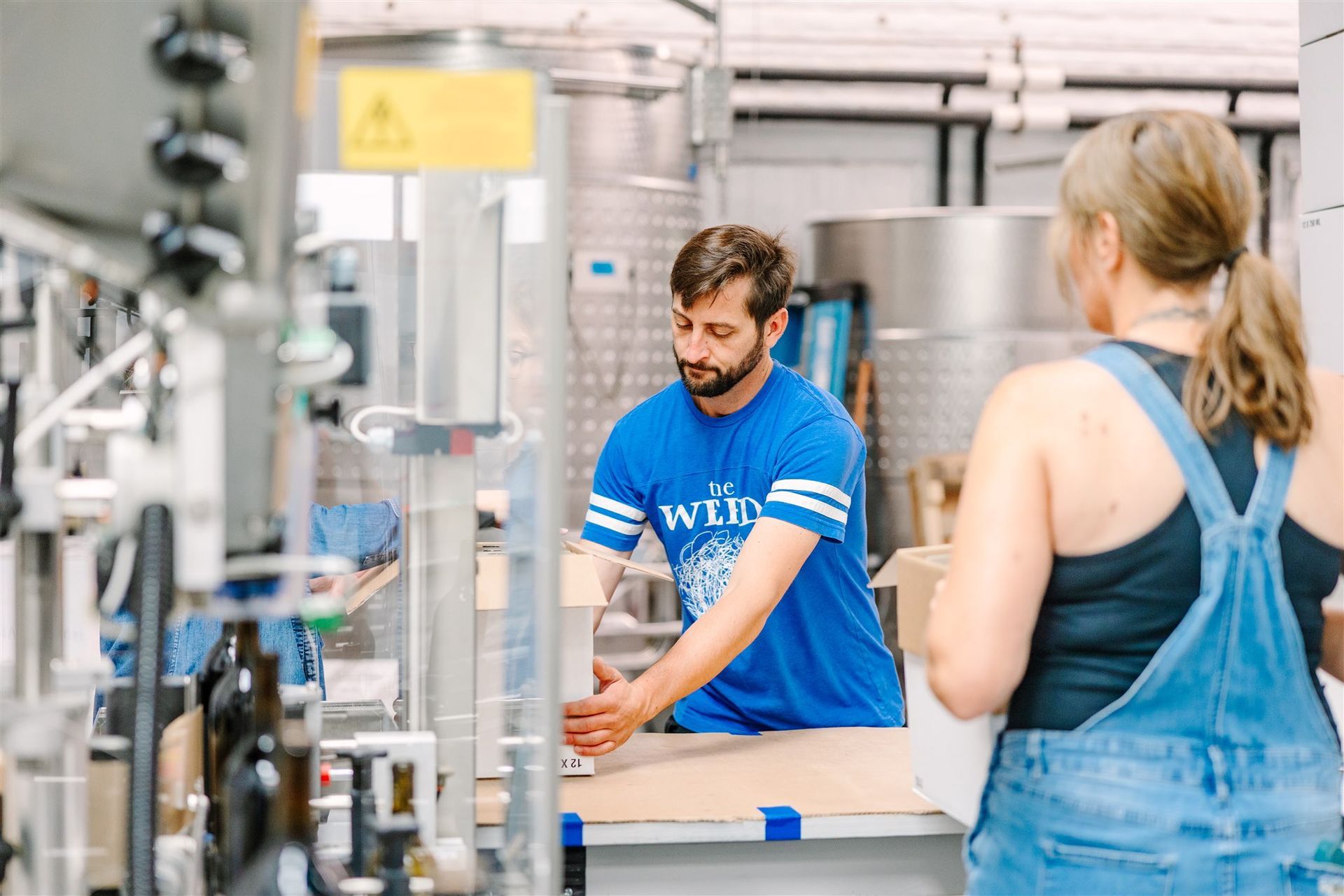 A man in a blue shirt is standing next to a woman in overalls in a factory.