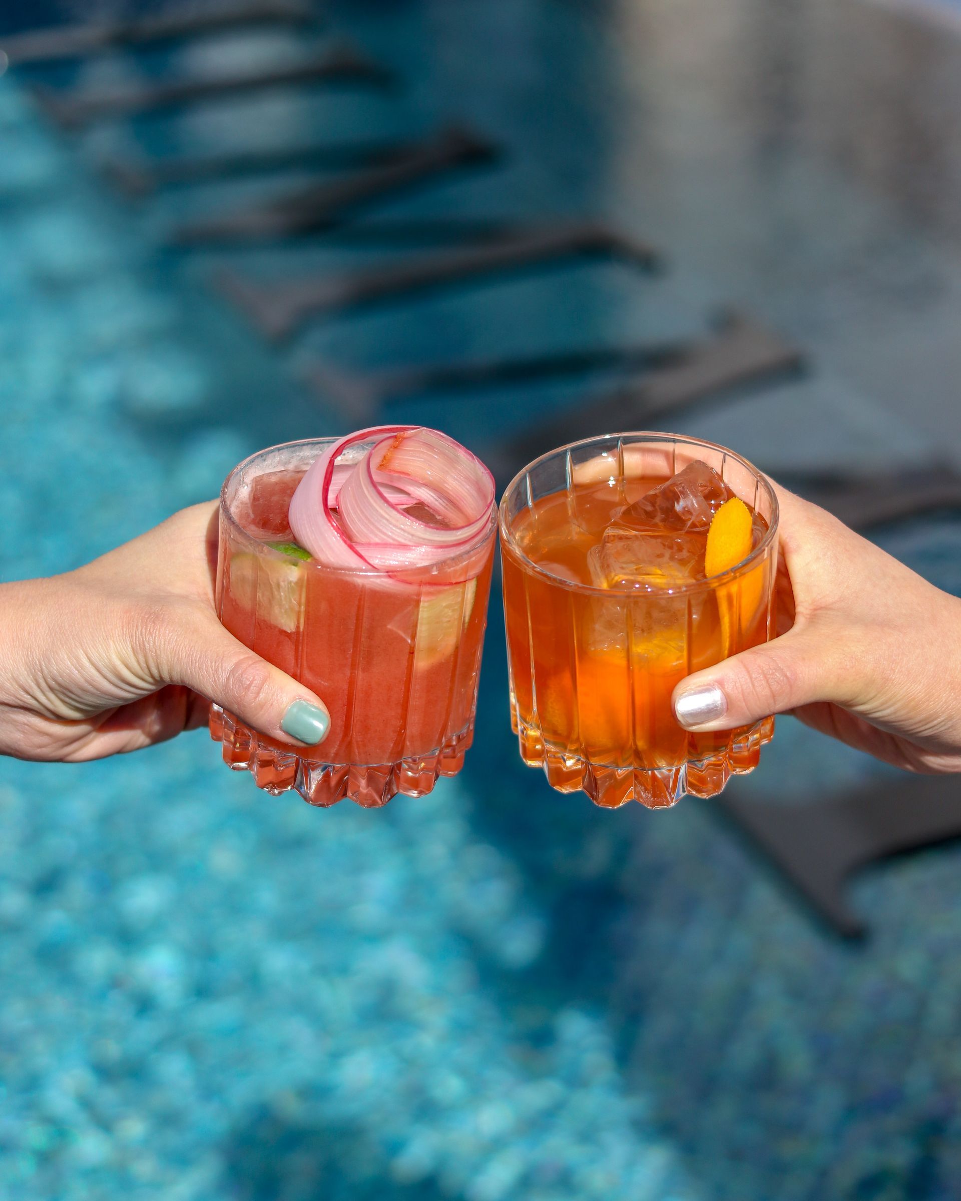 Two people are toasting with drinks in front of a pool.