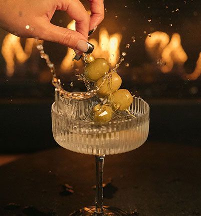 A person is pouring olives into a martini glass